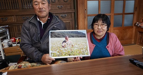 蕎麦畑での写真を持つ長谷川健一さん花子さん夫妻